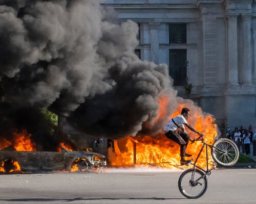 A person performs a bicycle wheelie on a street in front pf Philadelphia City Hall while large fires burn in the background, creating a dramatic contrast between the calm cyclist and the chaotic scene of flames and smoke.