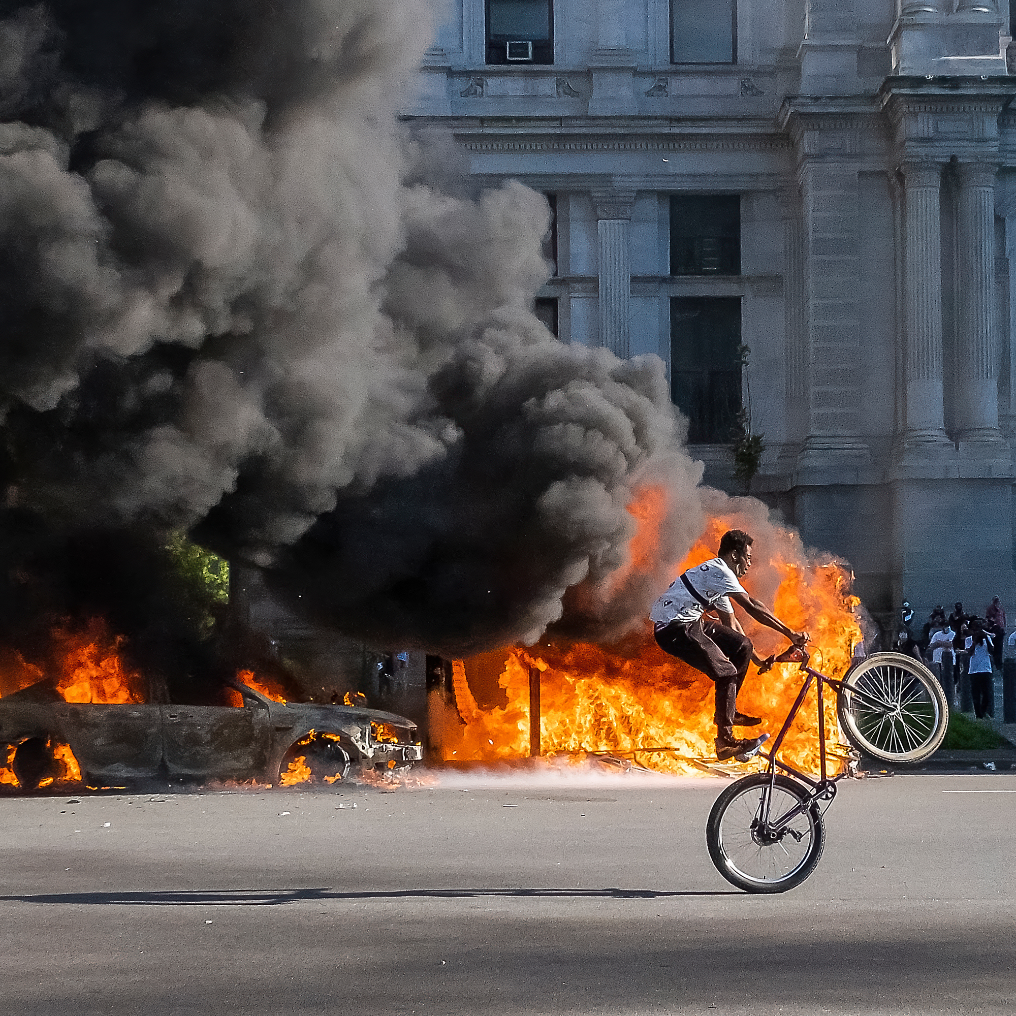 A person performs a bicycle wheelie on a street in front pf Philadelphia City Hall while large fires burn in the background, creating a dramatic contrast between the calm cyclist and the chaotic scene of flames and smoke.