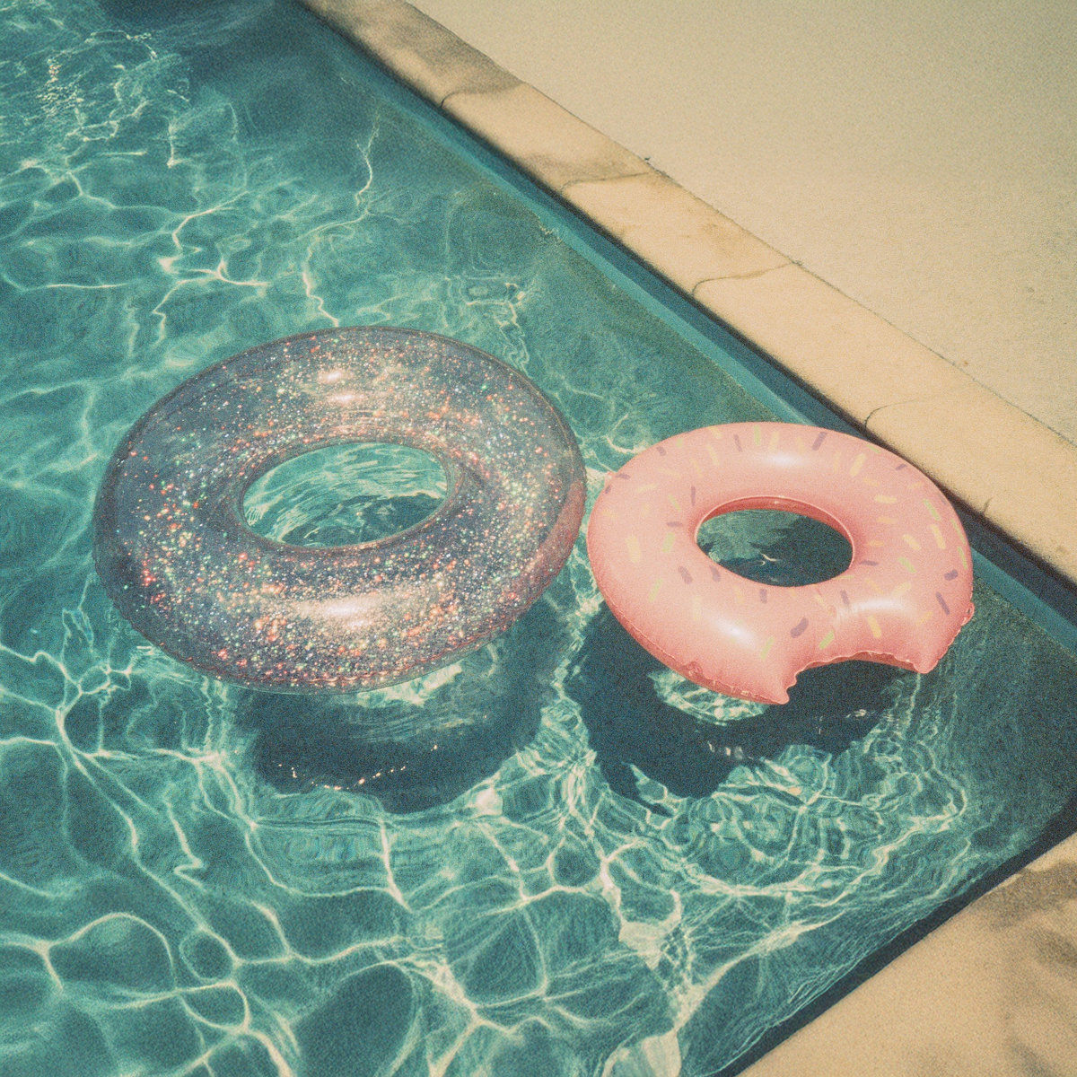 Two inflatable pool floats float on the surface of a clear, turquoise swimming pool. One is a transparent glittery ring, and the other is a pink donut-shaped float with colorful sprinkles and a bite mark design. Sunlight reflects off the water, creating rippling patterns. The pool is bordered by tan stone tiles.