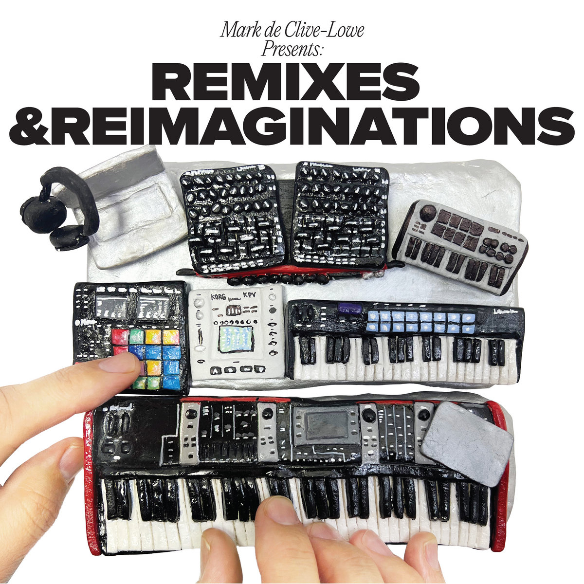 A miniature replica of Mark de Clive-Lowe's studio gear, seemingly hand-crafted from clay and painted in detailed color. Effects pads, MIDI keyboards, and controllers are arranged on a flat surface. The photo is taken from overhead, showing what appear to be Mark’s hands resting on the devices, as if mid-performance.